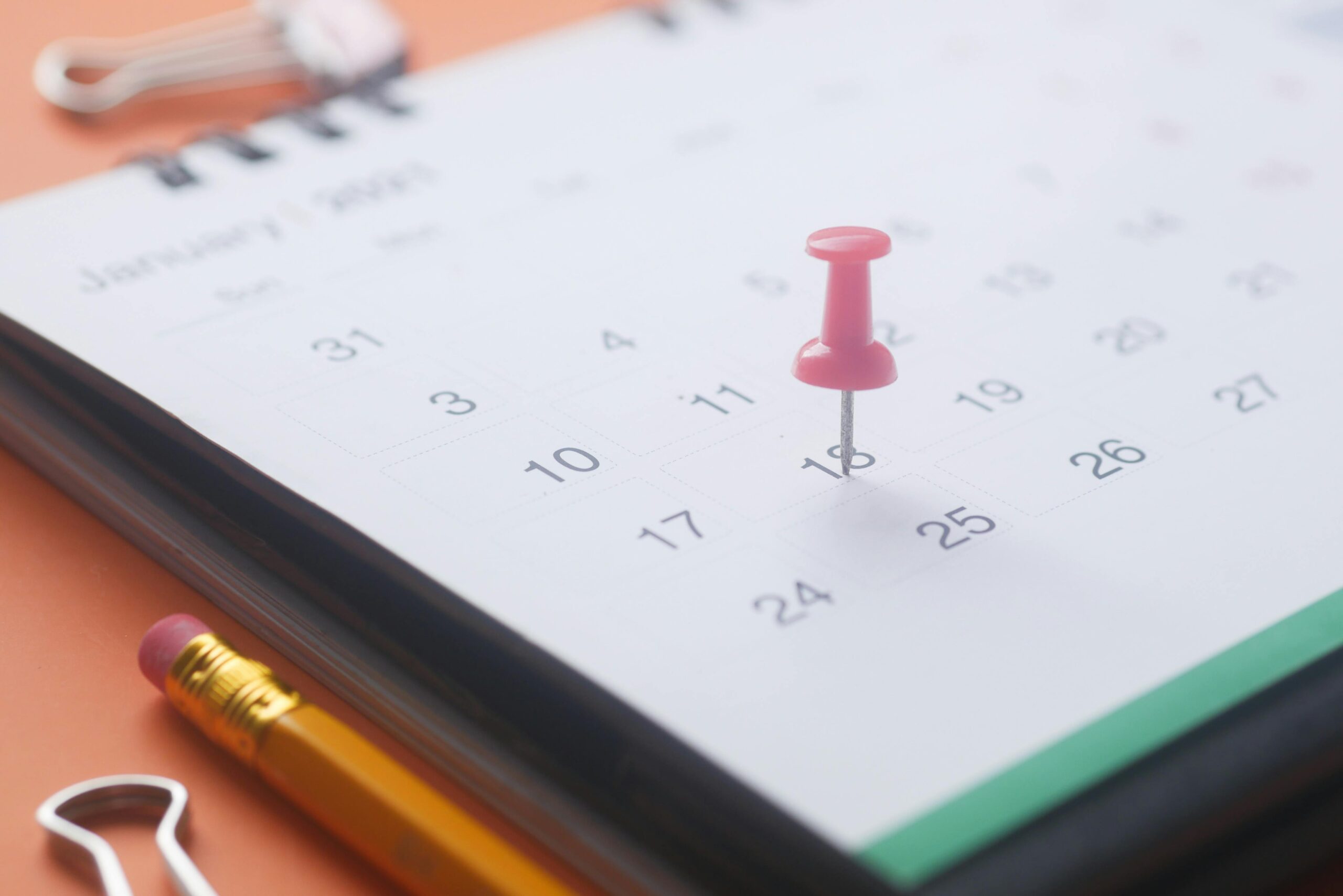 Booking Management A close-up of a calendar page with a pink push pin, pencil, and binder clip on an orange background.